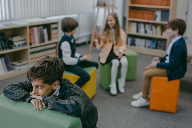 Drie kinderen spelen op de achtergrond van de foto een spelletje. Op de voorgrond kijkt een kind dat buitengesloten lijkt te worden sip voor zich uit.